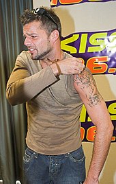 Ricky Martin looking to his right, while pulling up his the left sleeve of his t-shirt to reveal a tattoo.