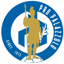 A blue circle with the text "Pro Palozzolo" "Since 1913" inside of the circle. Further into the circle is another white circle containing a blue design in the middle. There is a golden silhouette of a Roman soldier in front of everything.