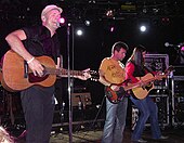 A man wearing a hat while playing a guitar and singing into a microphone. There is also a bassist and another guitarist to the right on stage