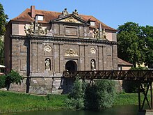 Photographie d'un bâtiment en pierre situé au bord d'un fleuve avec une façade décorée de six statues qui entourent une arche reliée à une passerelle en bois qui passe au-dessus de l'eau.