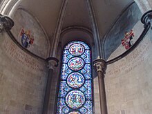 A semi-circular area with stone walls, a domed ceiling and a tall stained glass window with prominent circular decorations.