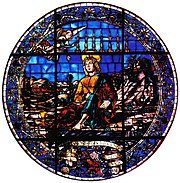 A circular stained glass window showing a saint with an angel and 7 candles above him. At his foot there is a golden chalice. There is a decorative frieze of flowers and oranges.