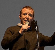 Guy Ritchie in a black shirt speaking while holding a microphone.