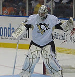 Hockey player in white uniform and goaltender's gear. He stands in front of the goal, legs shoulder length apart, and has raised his right arm as if to catch a puck, while his left arm holds a hockey stick.