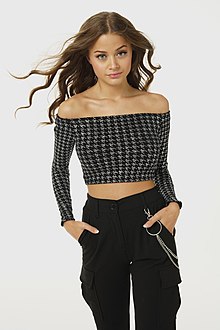 A young woman wearing a dark patterned crop top and black pants staring straight at the camera