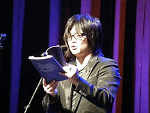 Standing Japanese man reading a book onstage at a microphone