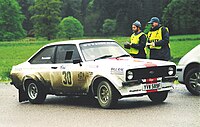 A Mark II rally car at a stage rally time control, displaying the extended wheel arches
