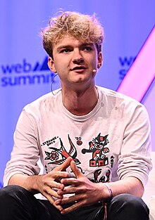 Casually dressed young man with curly hair talking while sitting on a chair.