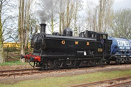 A pannier tank locomotive is pulling a milk wagon. The locomotive is painted black. The words "GREAT WESTERN" is shown in yellow with red shading on the side of the pannier tank. The front buffer beam is painted red.