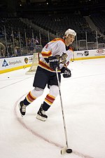 A hockey player skates around the net during warm-ups. He has one hand on his stick while he is skating with the puck on his back-hand. He is wearing a "traditional " Panthers' white jersey with a white helmet, white hockey socks with yellow and red horizontal stripes, black gloves, shorts and skates.