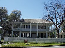 A gambrel-roofed house with large windows, wraparound balcony and first floor porch with Grecian columns. Sashed windows are framed by shutters.