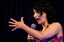 A surprised-looking woman with black curly hair extending one arm while holding a microphone with the other.