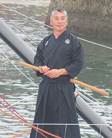 Debout sur le pont de son bateau, le navigateur est en tenue noire d'arts martiaux, un bâton passé à sa ceinture.