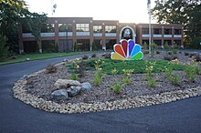 A two-story office building featuring the WYFF logo. In front is a turnout and driveway with a large landscaped element featuring a three-dimensional NBC peacock.