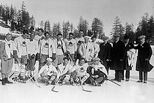 Black and white photo of a hockey team outside on natural ice, including fourteen players dressed in hockey equipment white sweaters with a maple leaf crest, and four men dressed in dark suits and overcoats
