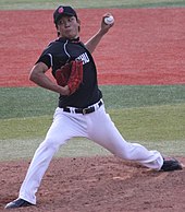 Yūdai Ōno wearing a navy baseball uniform and navy baseball cap in the process of pitching a baseball