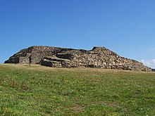 Vue de trois-quarts de l'ensemble du cairn, proue en premier plan. À la proue, la série de grosses dalles verticales du parement médian. Sur l'arrière du cairn, la partie détruite montre en coupe l'étagement des trois parements.