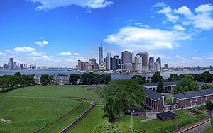 Aerial view of Governors Island in New York City on a sunny day. There is a lawn and some houses in the foreground, and skyscrapers in the background.