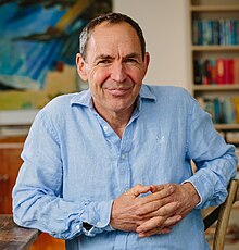 A man smiling, in a blue cotton shirt with a bookcase behind him.