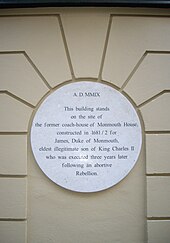 A white plaque on a beige wall noting where Monmouth House stood in Soho Square