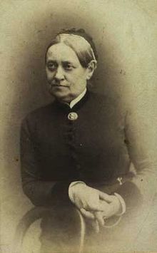 Black and white photo of a woman leaning with her right forearm on the back of a wooden chair