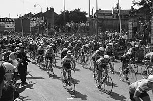 A black and white photograph of a large group of cyclists riding under a starting banner.