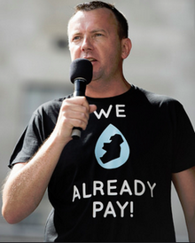 Man holding a microphone wearing a tshirt with a small map of Ireland and the phrase "We already pay"