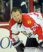 An ice hockey player stands bent at the waist with his stick horizontally across his knees. He wears no helmet and has a "spike" hair cut and goatee he is looking off to his left. He is wearing a "traditional" Panthers white jersey, the mostly white jersey has red shoulders which turn into a stripe that continues down the sleeves. The shoulders also have a patch on top which is a palm tree crossing a hockey stick in an "X" shape, there is a stylized sun as a background. The crest of the jersey has a stylized brown panther leaping at the viewer, to look as if the animal is attacking.