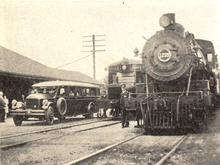 An old bus, a small railcar, and a steam locomotive at a railway station