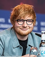 A red-haired man wearing glasses, a black shirt and a light blue jacket sits on a conference table