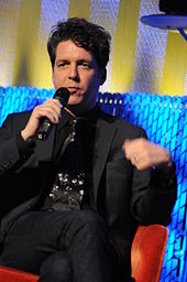 Joe Henry sitting on a sofa in a suit and talking to a microphone in his right hand.