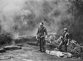 BW photo of two soldiers waiting in the jungle, a bodybag with a dead comrade at their feet
