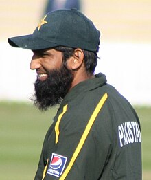 The head and shoulders shot of a beard man, wearing a dark green cricket cap and shirt.