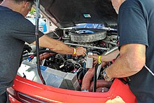 A V8 motor with a carburetor on it. There are two men on each side of the engine working on it.