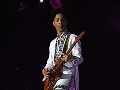 Prince playing the guitar wearing a white, jeweled shirt.