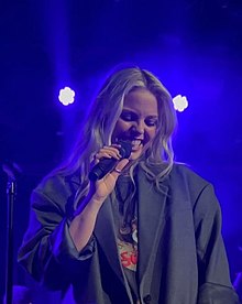 Profile picture of a White, blonde woman looking down, holding a microphone, and smiling