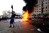 A rioter wearing a Canucks jersey cheers while a car burns, surrounded by a large crowd.