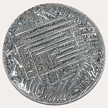 A shiny silver-white medallion with a striated surface, irregular around the outside, with a square spiral-like pattern in the middle