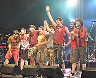 Eight performers stand at the front of a stage, and one holds their arm in the air