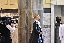 on the left a TV camera, in the centre a smiling Sandra McKie in ceremonial robes with a staff over her shoulder, on the right a bust of Kate Shepherd. The building is marble and timber.