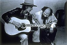 Bob Dylan playing an acoustic guitar as Allen Ginsberg, seated to his left, pays close attention to his fingering.
