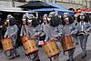A group of drummers wearing costumes to look like dogs wearing prison dress, playing as they walk along a narrow street