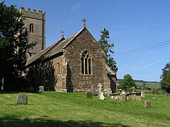 Stone building with square tower, partially obscured by trees. In the foreground are gravestones.