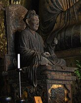 Three-quarter view of a cross-legged seated statue placed on a throne. His right arm rests on his leg, the left arm is bend and slightly raised. Color photograph.