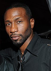 Leon Robinson with short cropped beard and mustache. He is wearing a black shirt and shiny, black tie.