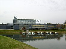 Photographie du Jardin du Monde, montrant un bâtiment bas incurvé, à structure de métal et panneaux pleins de couleur gris foncé, avec en son centre une serre enserrée dans un réseau métallique. Devant s’étale, entouré de pelouses, un plan d’eau avec un ponton et des petits bateaux à quai.