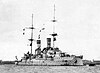 A large, light gray battleship bristling with guns sits in harbor