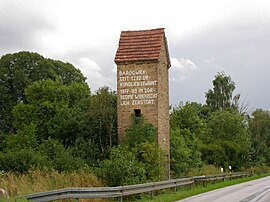 Une petite tour électrique en brique avec un toit en tuiles rouges se trouve à côté d'une route et devant une forêt à l'arrière-plan. Il y a une porte en bois sur la droite et une fenêtre au premier étage. Le mur du second étage porte une inscription blanche peinte à la main : « BARDOWIEK: SEIT 1292 URKUNDLICH ERWÄHNT 1977-'89 IM "DDR"-REGIME WIDERRECHTLICH ZERSTÖRT. »