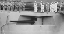 photograph of a cannon mounted in an armored bulge in the side of a ship, just below a deck with a number of sailors.