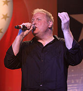 A blond-haired man wearing a black shirt, singing into a microphone